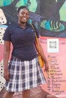 Smiling woman wearing a navy pinstripe polo shirt and plaid pleated skirt, carrying a yellow shoulder bag, standing in front of a colorful mural wall.