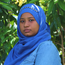 Picture of Suhayla Portrait - she is facing the camera and is wear a blue Jean jacket and blue scarf and she has green leaves in her backdrop.